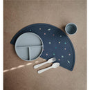 Mushie silicone placemat in dark blue with planet designs, featuring a gray divided plate, white utensils, and a cup.
