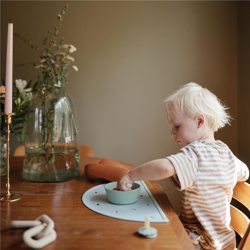 Child using a blue Mushie silicone placemat with a bowl, surrounded by a candle and flowers on a wooden table.