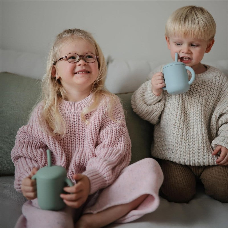 Two children sit on a couch, smiling and holding colorful mugs with silicone straws, dressed in cozy sweaters.