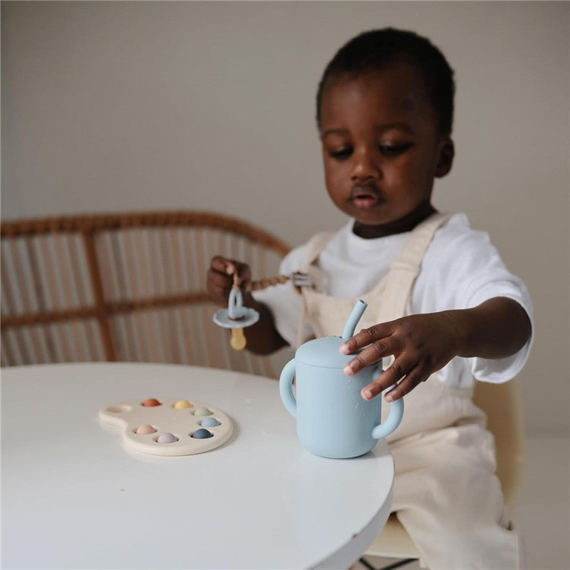 A young child wearing a white shirt and overalls holds a blue silicone cup with a straw, while a pacifier hangs from their other hand. A paint palette with pastel colors is on the table.