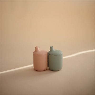 Silicone sippy cups in blush and green colors, designed for toddlers, resting on a light background.