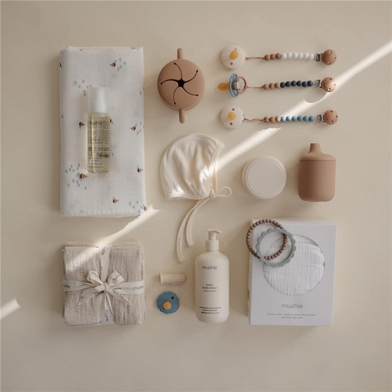 Mushie Silicone Sippy Cup in natural color, surrounded by baby accessories including a muslin blanket, body lotion, and teething toys.