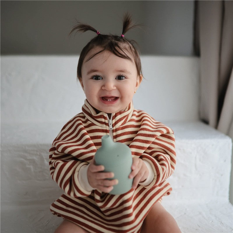 Smiling baby wearing a striped outfit, holding a powder blue Mushie silicone sippy cup.