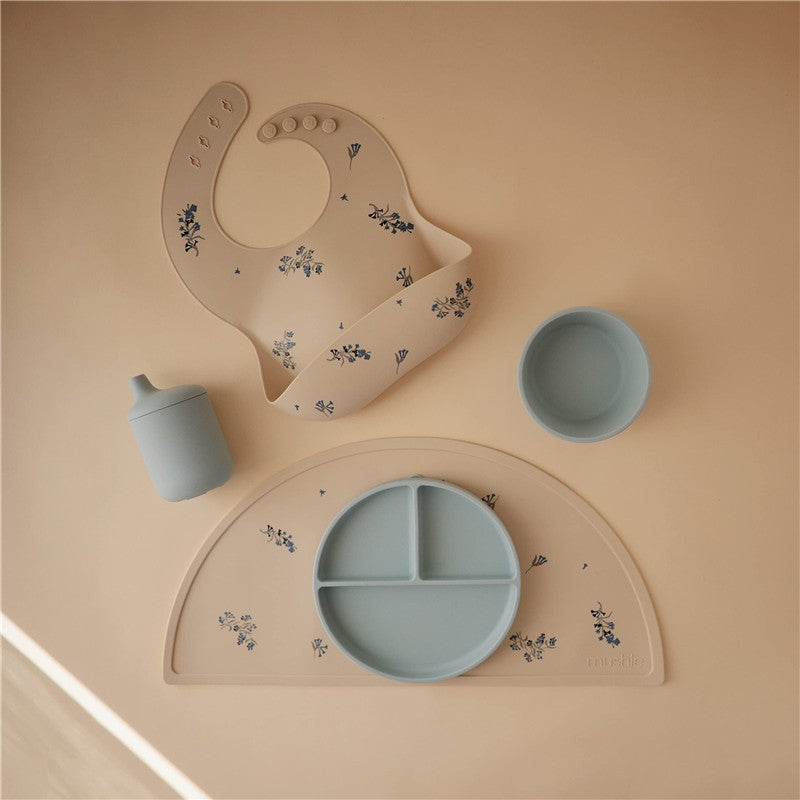 Mushie Silicone Sippy Cup in powder blue, with a matching plate, bowl, and bib, all featuring floral designs on a neutral background.