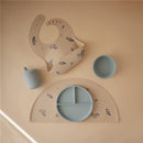 Mushie Silicone Sippy Cup in powder blue, with a matching plate, bowl, and bib, all featuring floral designs on a neutral background.