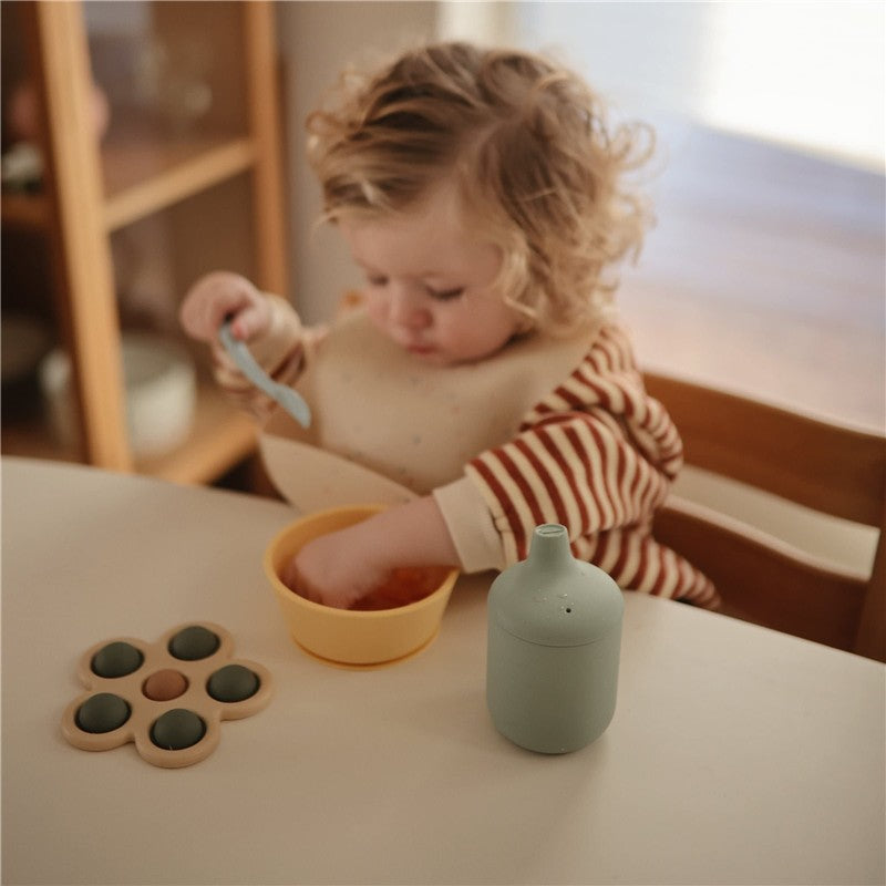 Child with a striped shirt eating from a yellow bowl while a powder blue Mushie Silicone Sippy Cup sits on the table.