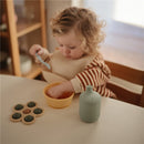 Child with a striped shirt eating from a yellow bowl while a powder blue Mushie Silicone Sippy Cup sits on the table.