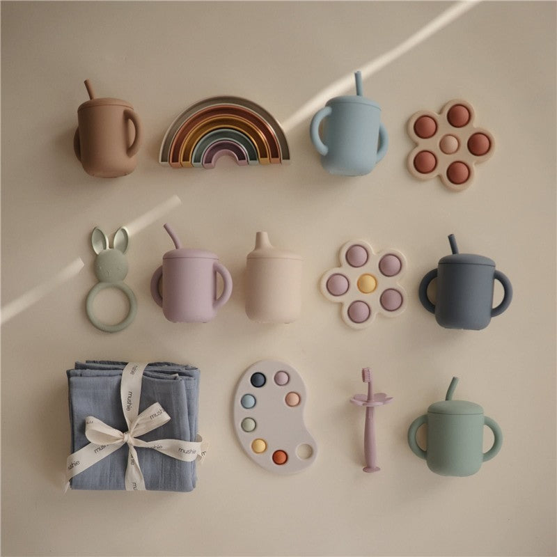 Mushie Silicone Sippy Cups in various colors, accompanied by playful accessories like a rainbow, silicone teething ring, and a blue cloth.