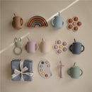 Mushie Silicone Sippy Cups in various colors, accompanied by playful accessories like a rainbow, silicone teething ring, and a blue cloth.