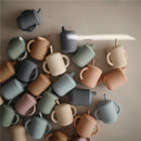 Silicone sippy cups in various pastel colors, including powder blue, are scattered on a light surface.