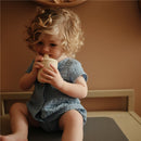 Child with curly hair sitting on a surface, drinking from a Mushie Silicone Sippy Cup in Shifting Sand color.