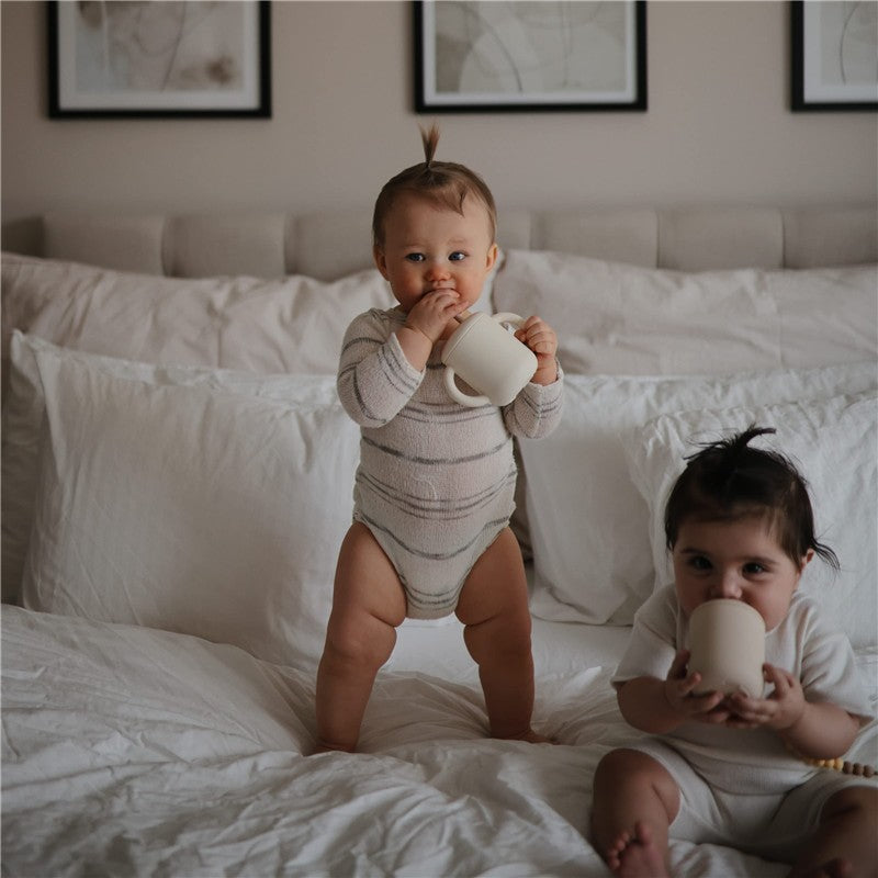 Two toddlers sit on a bed holding Mushie Silicone Sippy Cups in shifting sand color. One child stands, sipping from the cup.