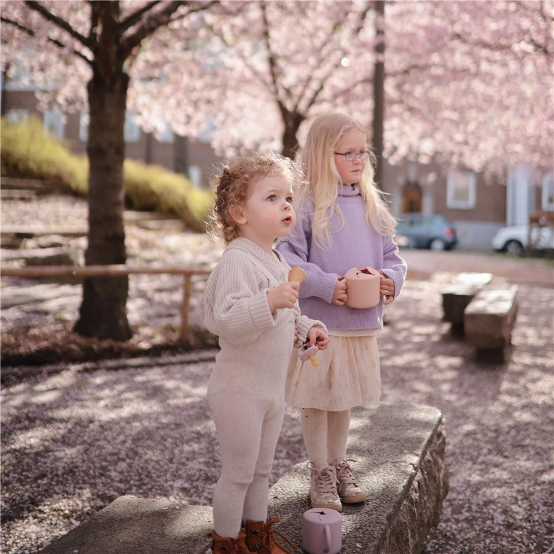 Two young children stand outdoors under cherry blossom trees, one holding a Blush Mushie Silicone Snack Cup and a cookie.