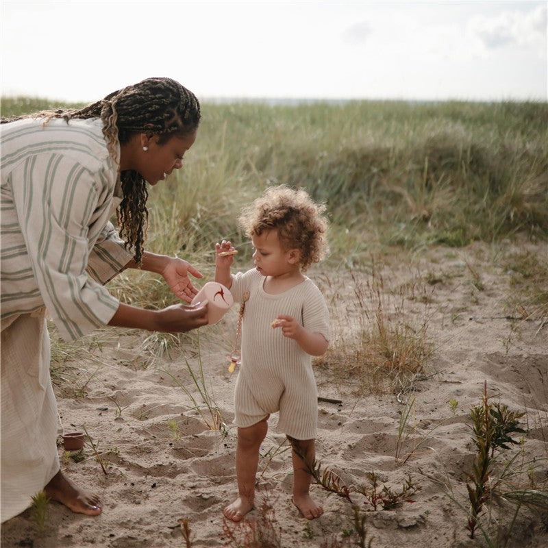 A child in a beige outfit reaches for a blush Mushie silicone snack cup held by an adult, set in a grassy outdoor scene.