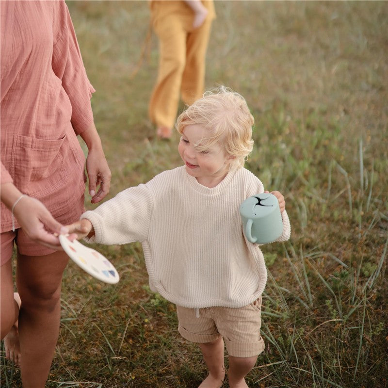 Child in a light sweater holds a Cambridge Blue Mushie Silicone Snack Cup, reaching for a plate from an adult.