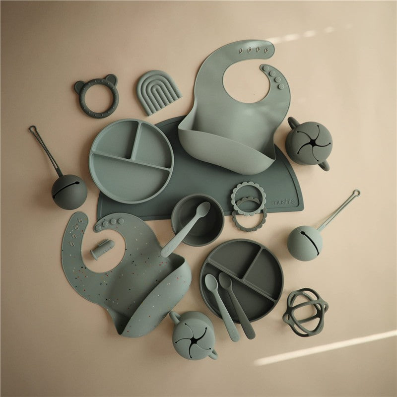 Silicone snack cup in Cambridge Blue surrounded by various silicone feeding accessories including bibs, plates, and utensils.