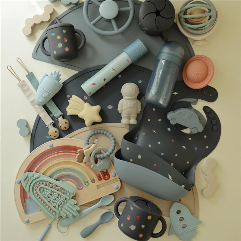Mushie Silicone Snack Cup in a planets design, surrounded by playful silicone toys, cutlery, and a colorful rainbow mat.
