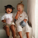 Two children sit on steps, holding Mushie silicone snack cups in pink and blue, smiling joyfully.