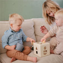 Children playing with a shape sorter toy on a couch; a boy holds a cup while another child places shapes into the toy.