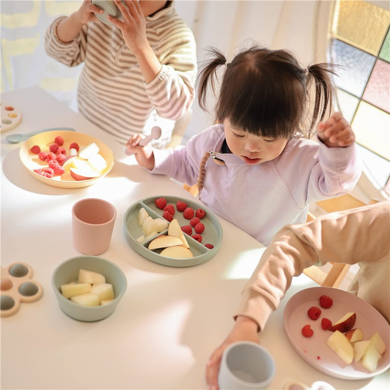 Two children enjoying snacks at a table with silicone suction bowls, featuring fruit like apples and raspberries.