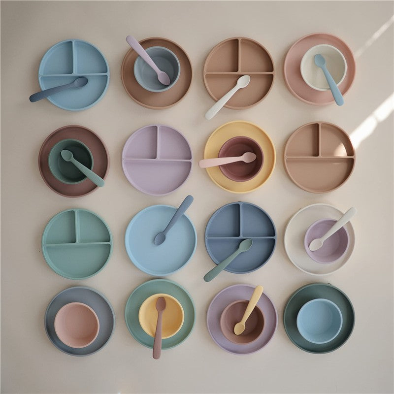 Mushie Silicone Suction Bowl in Cambridge Blue among a variety of colorful silicone plates and spoons arranged artfully.