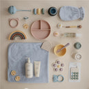 Mushie silicone suction plate in blush color, surrounded by baby products like utensils, bath items, and toys on a light surface.