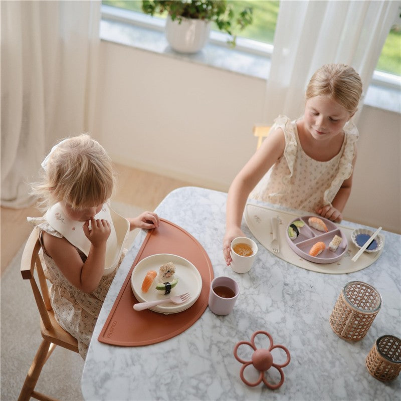 Two young girls are seated at a marble table, one with a Mushie silicone suction plate in ivory, while the other serves sushi and drinks.