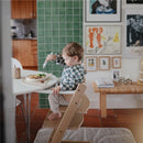 Child in a high chair using a fork to eat from a Mushie silicone suction plate in ivory, with a green tiled wall in the background.