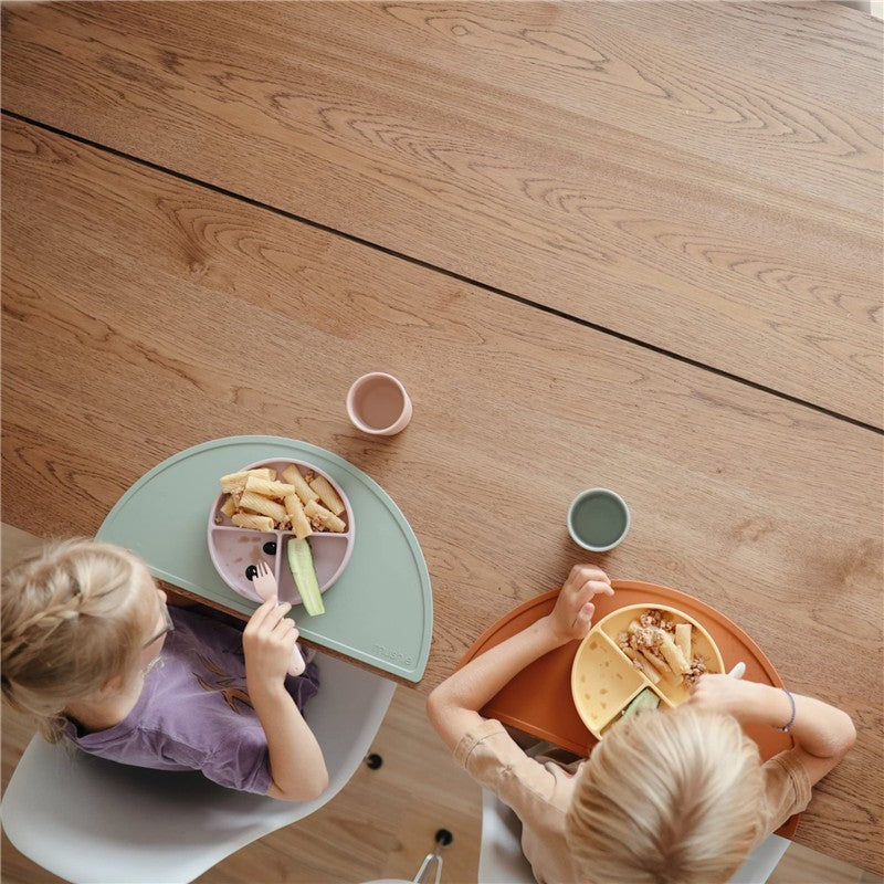 Two children eat from colorful silicone suction plates on a wooden table, featuring pasta, celery, and dipping sauces.