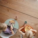 Two children eat from colorful silicone suction plates on a wooden table, featuring pasta, celery, and dipping sauces.