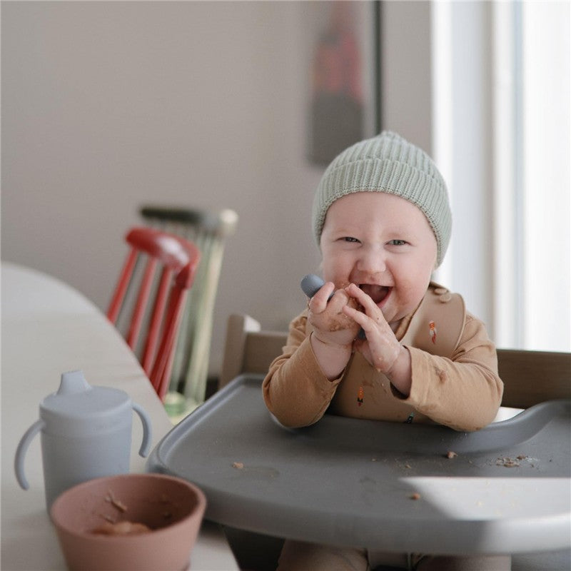 Toddler in a high chair, wearing a light green hat, happily plays with a silicone spoon, surrounded by food and cups.