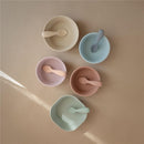 Silicone toddler starter spoons in natural and shifting sand colors, featured in various bowls on a neutral surface.