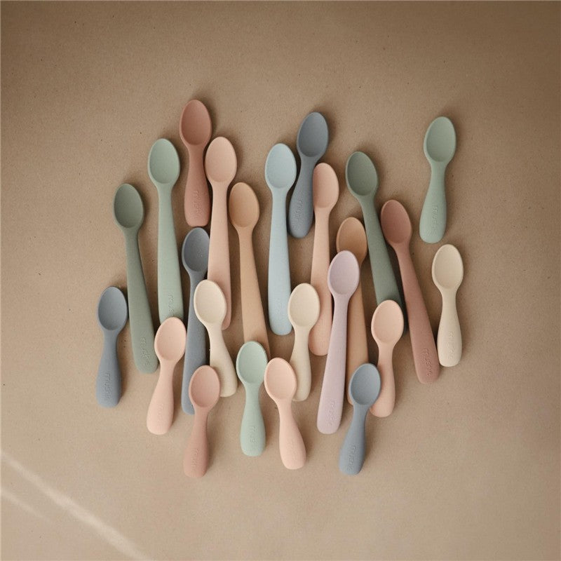Silicone toddler starter spoons in various soft colors, arranged on a neutral background.