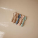 Silicone toddler starter spoons in natural and shifting sand colors, arranged on a neutral background.
