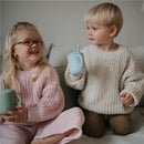 Two children seated on a sofa, each holding a silicone training cup. The boy has a powder blue cup with a straw, while the girl has a green cup. Both are smiling and wearing cozy sweaters.