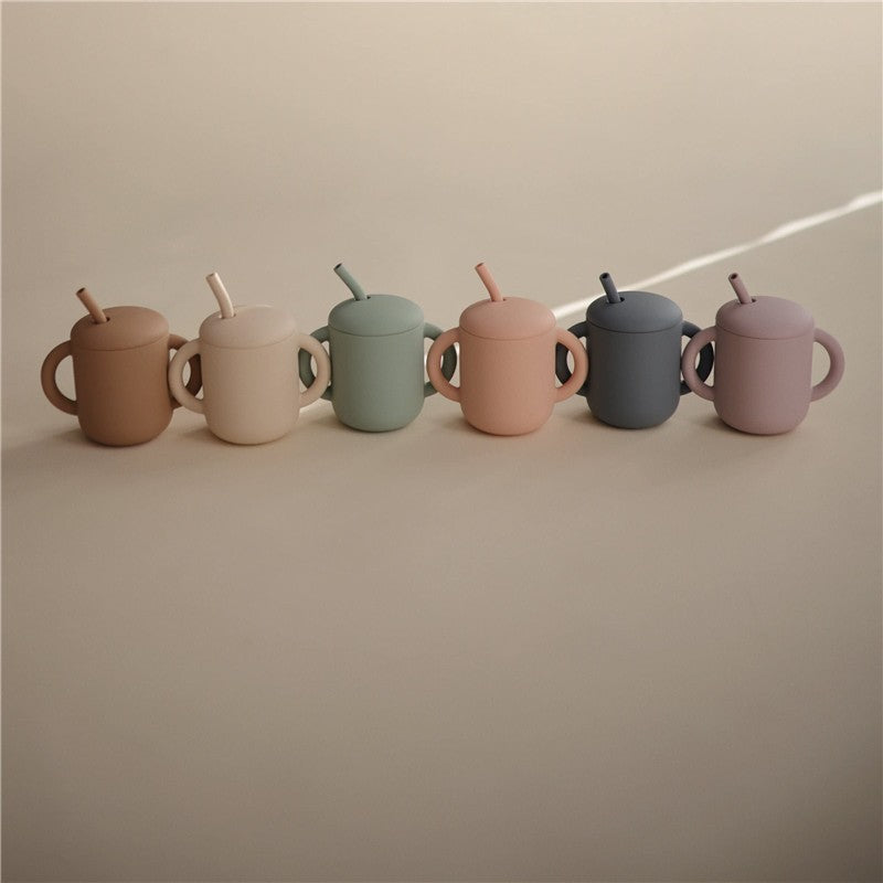 Silicone training cups in various pastel colors with straw, showcasing the Mushie brand, arranged in a row.