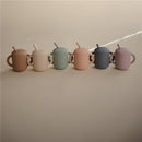 Silicone training cups in various pastel colors with straw, showcasing the Mushie brand, arranged in a row.