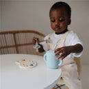 Mushie Silicone Training Cup in powder blue being used by a child, beside a color palette and pacifier on a table.