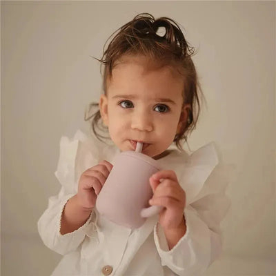 A child with dark hair holds a soft lilac silicone training cup with a straw, sipping from it.