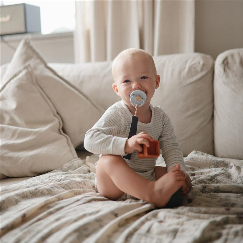 Baby sitting on a couch holding a brown toy, wearing a light gray outfit and a pacifier, surrounded by soft pillows.