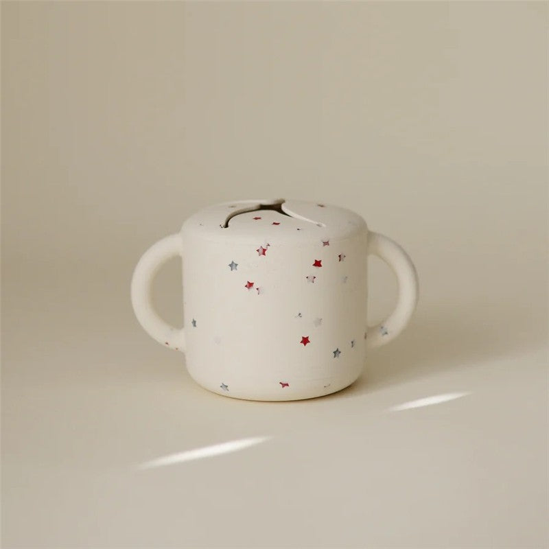 Mushie Snack Cup in white with red and blue stars, featuring two handles and a lid with a small opening.