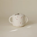 Mushie Snack Cup in white with red and blue stars, featuring two handles and a lid with a small opening.