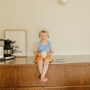 A young child wearing a green beanie and a blue tank top sits on a wooden counter, holding a Mushie Snack Cup in a light color, smiling.