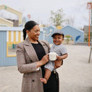 A woman smiles while holding a baby in a park, both enjoying the outdoor setting with colorful play structures in the background.
