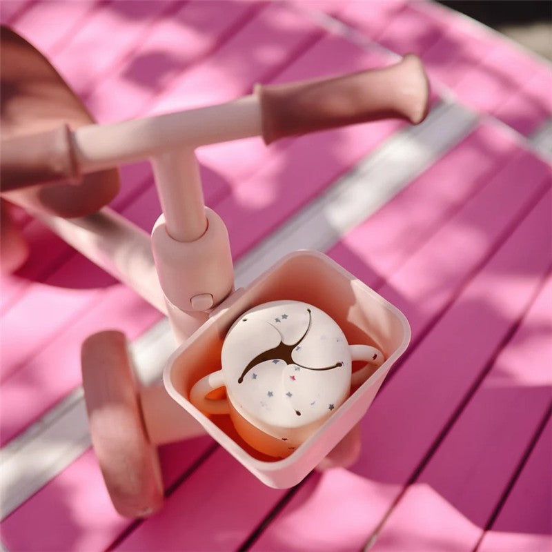 Mushie Snack Cup in soft pink with a star pattern, resting in a toy stroller on a vibrant pink surface.