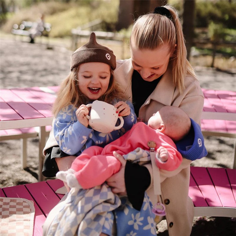 A smiling young girl in a blue sweater holds a white Mushie Snack Cup while sitting with a woman and a baby, all outdoors.