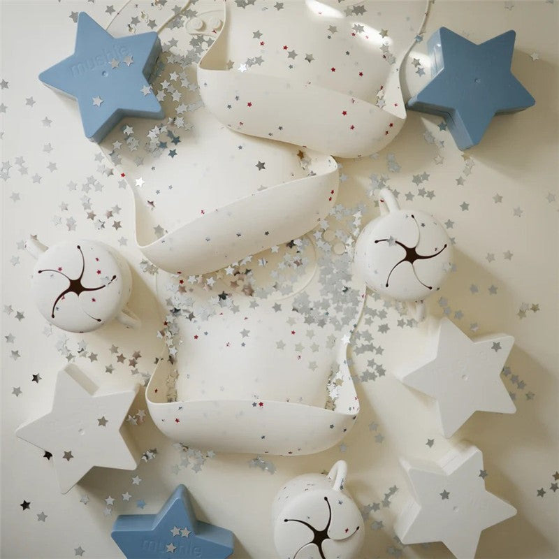 Mushie Snack Cup in white with star designs, surrounded by star-shaped confetti and a blue star-shaped object.