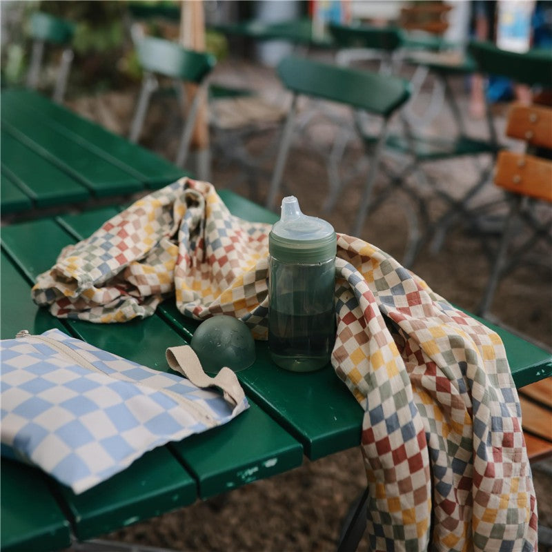 Mushie Soft Spout Sippy Cup in Dried Thyme resting on a green table, surrounded by a patterned cloth and a small ball.