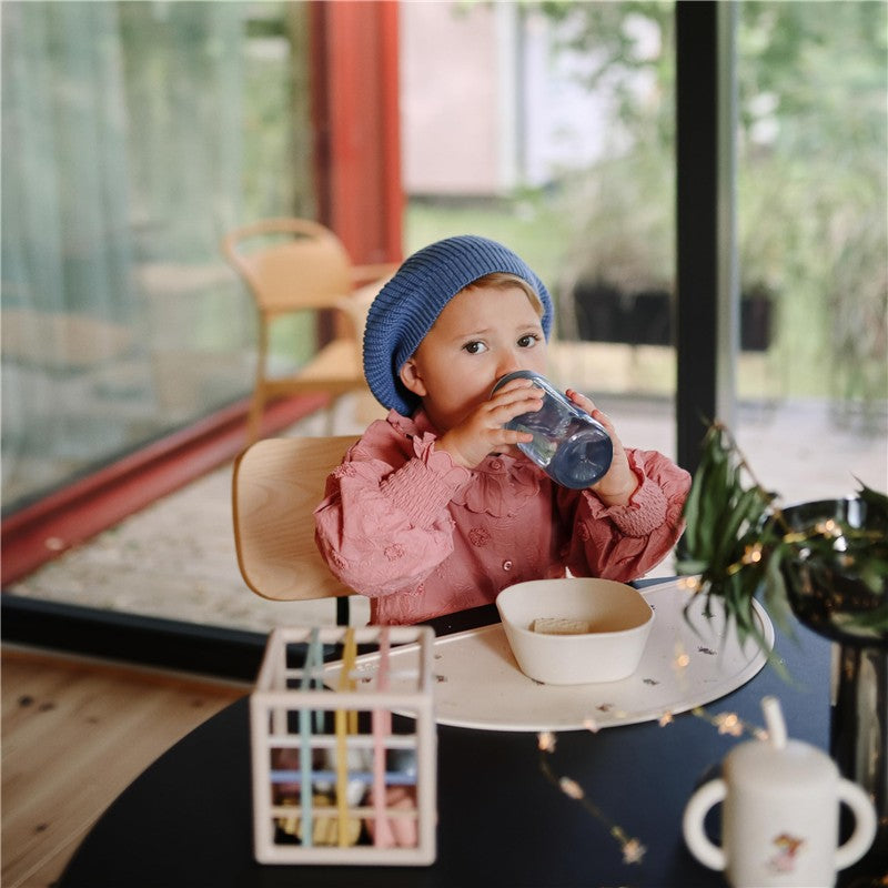 Child in a blue beanie drinking from a Mushie Soft Spout Sippy Cup - Tradewinds at a stylish table.