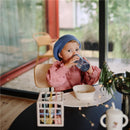 Child in a blue beanie drinking from a Mushie Soft Spout Sippy Cup - Tradewinds at a stylish table.